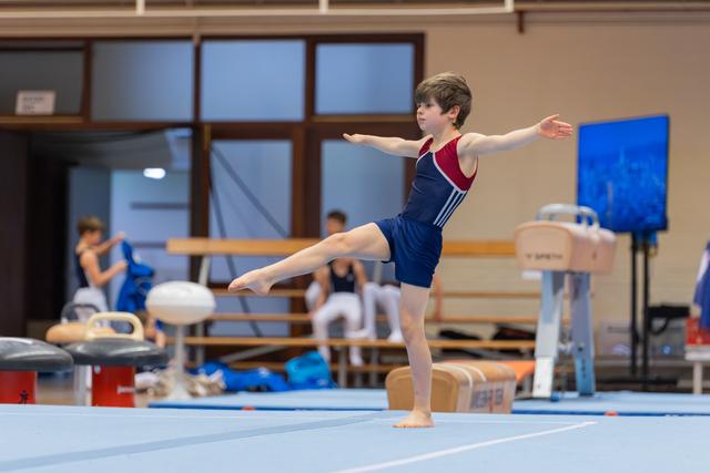 Young male gymnast balances on one leg, arms outstretched, focused expression during an indoor gymnastics event.