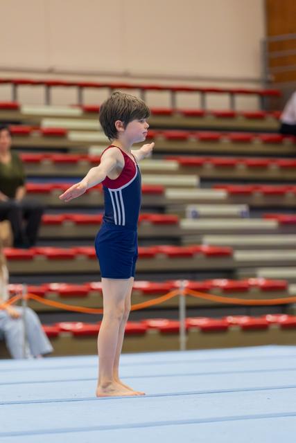 Young male gymnast stands barefoot on floor apparatus, arms extended, focused expression, bleachers visible behind.
