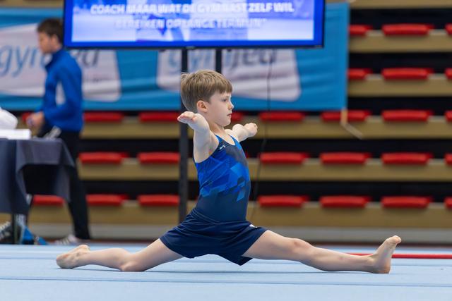 Young boy performs a straddle split on the floor exercise mat, arms forward, expression calm and concentrated.
