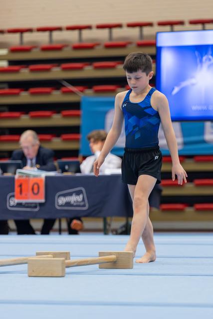 Focused young male gymnast walks barefoot on blue floor, studying wooden apparatus blocks, judges visible behind him.