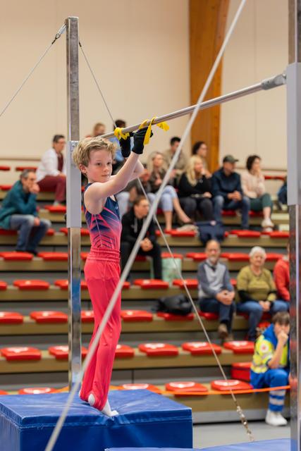 Focused young male gymnast grips the high bar with determination, standing on a blue mat block before his routine.
