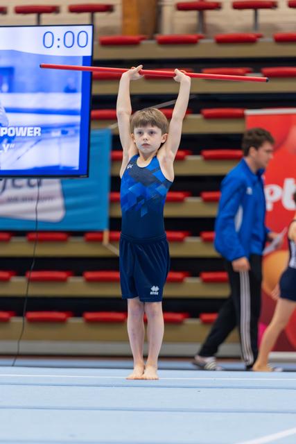 Young boy in blue gymnastics outfit holds a red bar overhead with focused expression on a competition floor.
