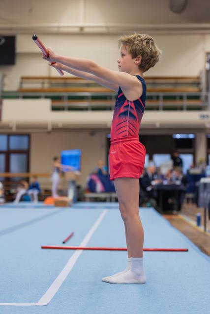Young gymnast in red and navy leotard stands proudly on blue mat, arms extended holding a baton, focused expression.