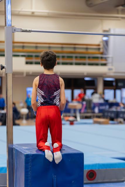 Young male gymnast stands on a blue block, back turned, gazing at the high bar ahead with quiet focus in a gym.
