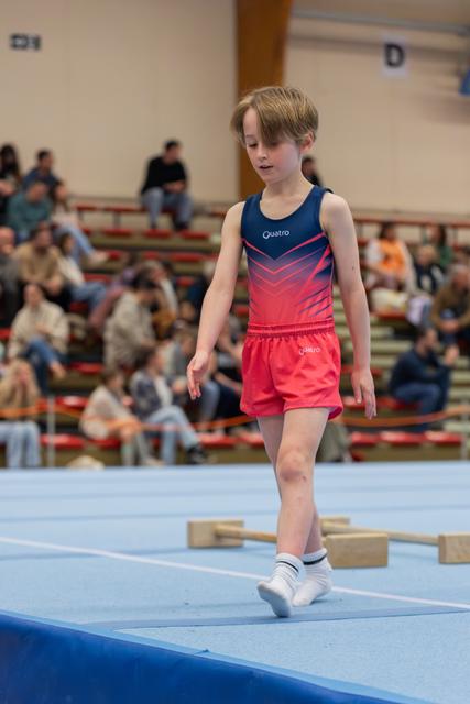 Focused young boy in red and navy Quatro leotard walks carefully across a blue gymnastics mat before a crowd.