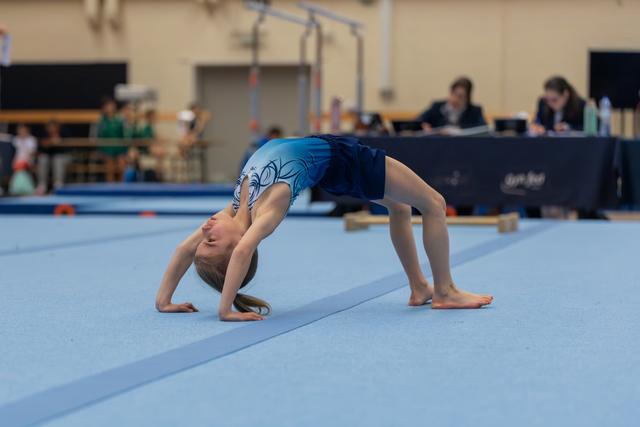 Young girl in blue leotard holds a bridge pose on the floor mat during a gymnastics meet, head tilted back.