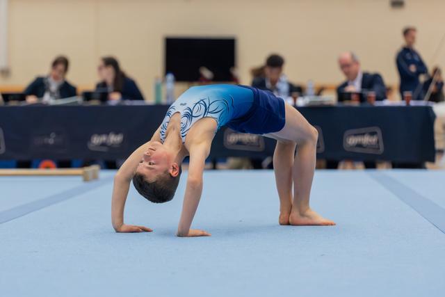 Young gymnast performs a back bridge on the blue floor mat, judges panel visible in the background.
