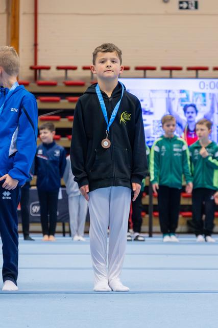 Young boy stands solemnly on the podium wearing a bronze medal and black NEIK hoodie at a gymnastics competition.
