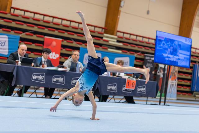 Young girl in blue leotard performs a one-handed cartwheel on floor exercise, judges panel visible behind her.
