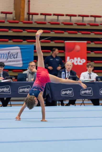 Young gymnast performs a cartwheel on the floor exercise mat, judges observing at the scoring table behind.