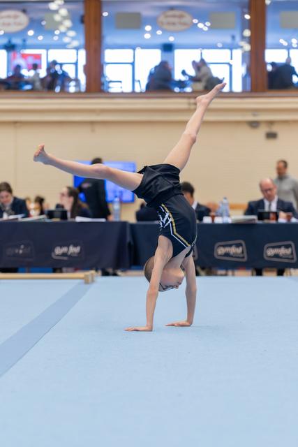 Young gymnast performs a one-handed cartwheel on the floor mat, legs split wide, at an indoor gymnastics meet.