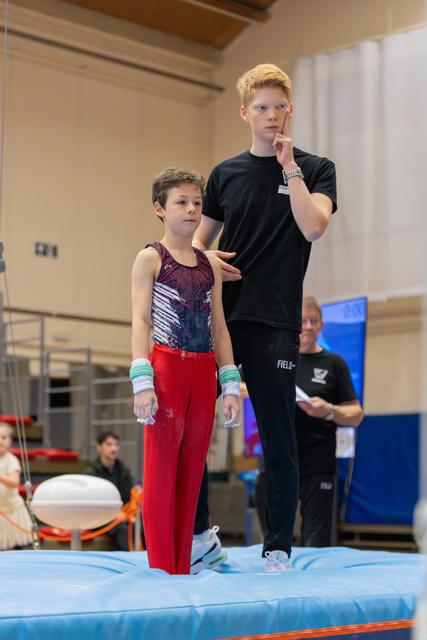 Young boy in red gymnastics attire stands attentively while an older red-haired teammate gestures and coaches him on a blue mat.