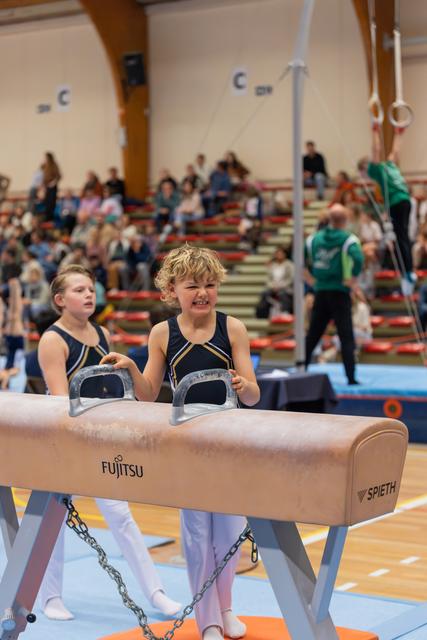 Young gymnast grips pommel horse handles with fierce determination, teammate watching behind, packed gymnasium crowd in background.