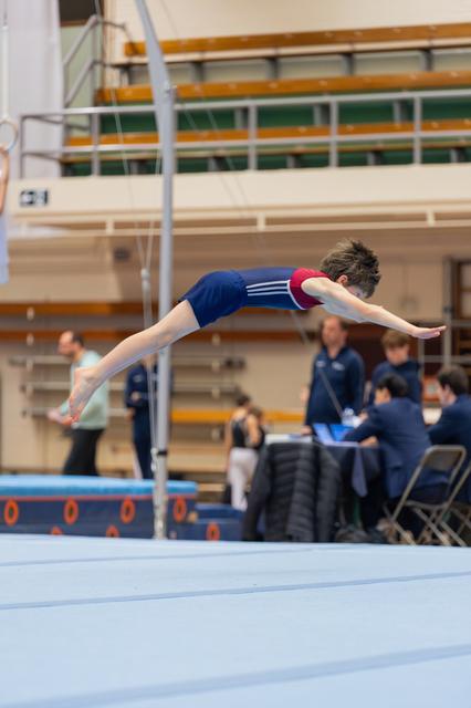 Young male gymnast soars horizontally, arms outstretched, body fully extended mid-air above a blue mat in an indoor gym.