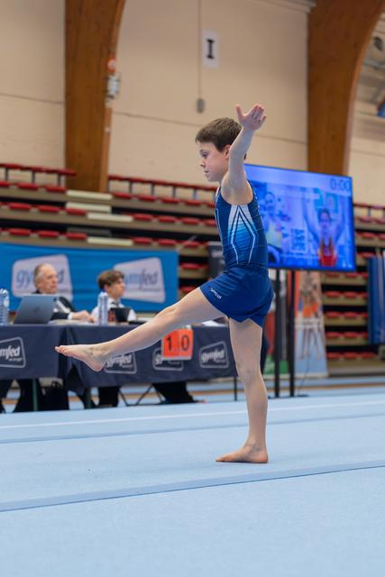 Young male gymnast performing a floor routine, balancing on one leg with arm raised, focused expression in a competition hall.