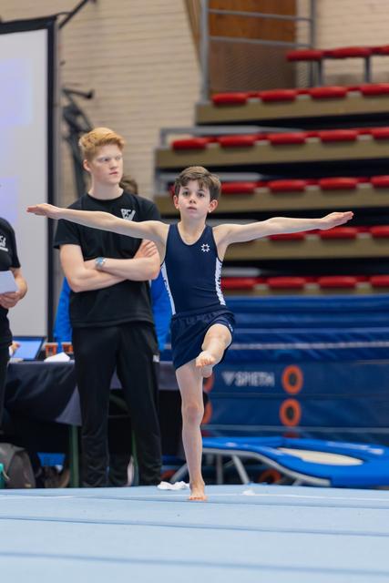Young boy gymnast balances on one leg, arms outstretched, focused expression during floor exercise while coach watches behind.