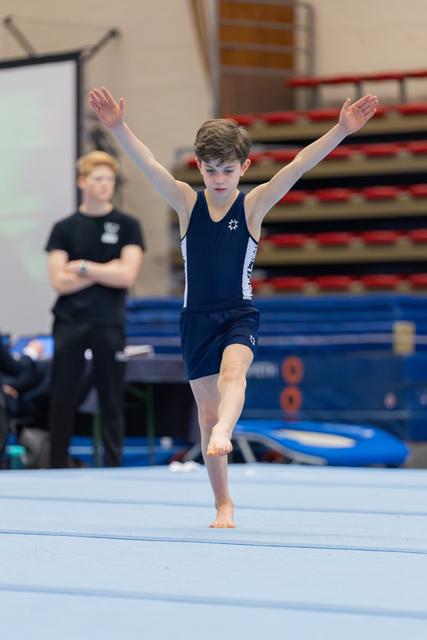Young male gymnast performs floor routine on one foot, arms raised, with focused expression. Coach watches in background.