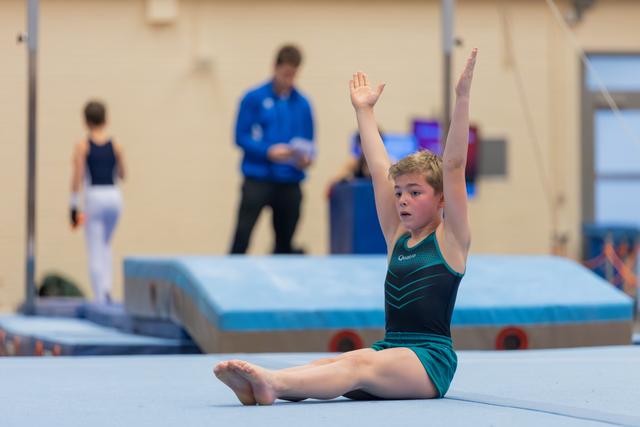 Young gymnast sits on the floor mat, arms raised triumphantly after completing a routine at an indoor gymnastics event.