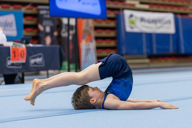 Young boy in blue leotard performs a backbend pike on the floor exercise mat during a gymnastics competition.