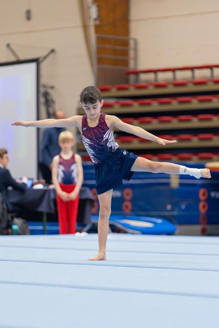 Young male gymnast balances on one leg with arms outstretched during a floor routine at an indoor gymnastics meet.