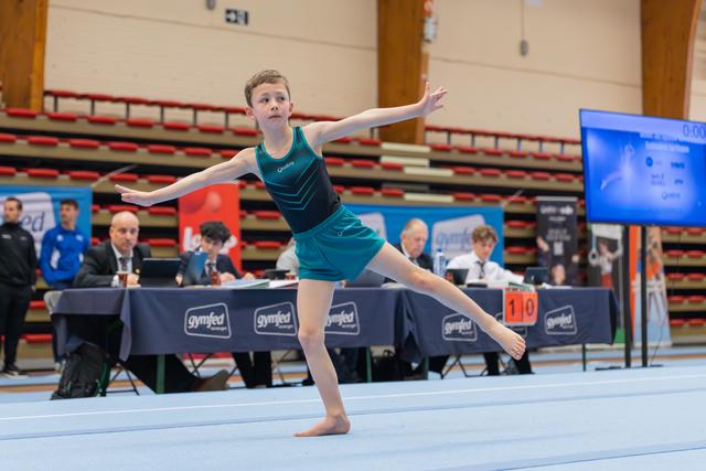 Young gymnast in teal leotard performs a graceful floor routine, arms extended, at a Gymfed judged event.