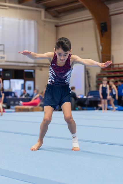 Young male gymnast performs floor routine, arms outstretched for balance, focused expression, ankle bandaged, in a gymnasium.