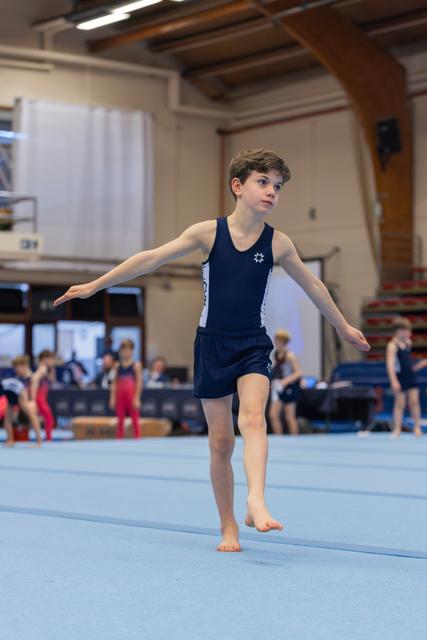 Young male gymnast balances on one foot on the floor mat, arms extended, focused expression in a busy gymnasium.