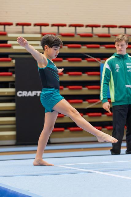 Young gymnast performs a focused floor routine, leg extended mid-step, while a coach in green watches attentively behind.