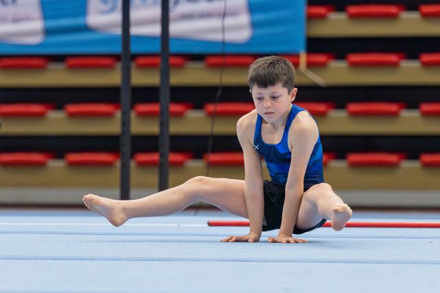 Focused young boy gymnast performing a floor split stretch, barefoot on a light blue mat in a sports hall.