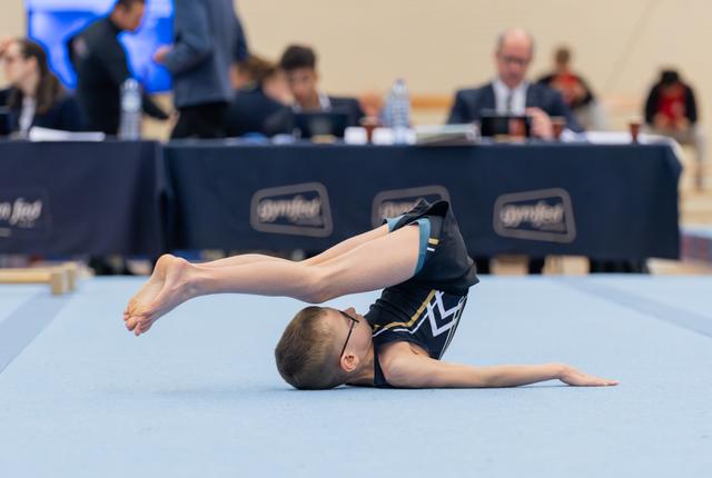 Young gymnast wearing glasses performs a floor exercise, lying face-down with legs raised high in a controlled tuck position.