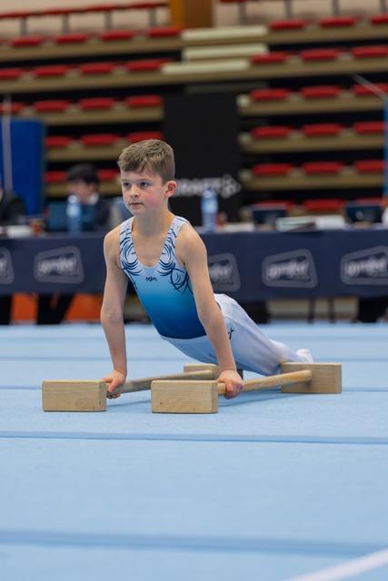 Young boy in blue leotard performs floor exercise on parallettes, focused expression, at indoor gymnastics event.