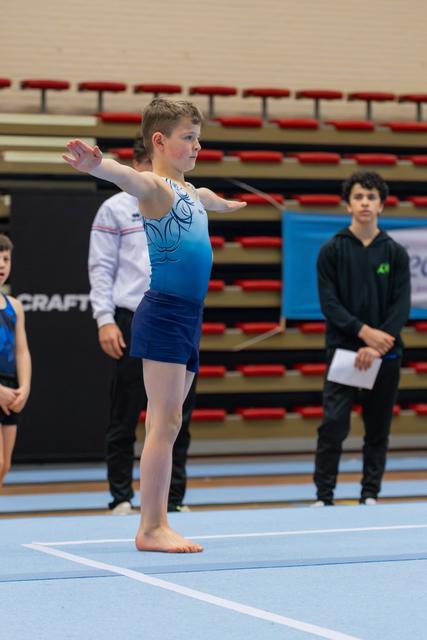 Young gymnast in blue leotard strikes a precise pose with arms extended on the floor mat, focused and composed.