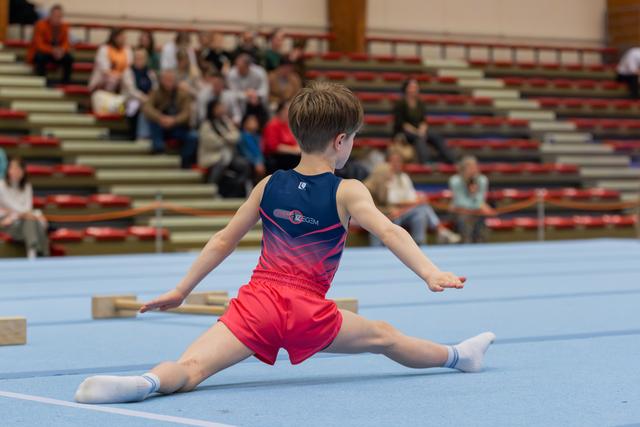 Young boy gymnast performs a splits on the blue floor mat, arms extended, focused during a gymnastics meet.