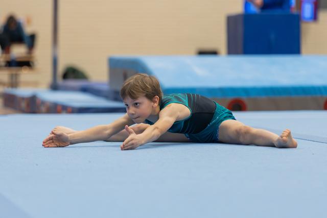 Young boy in teal leotard performs a straddle split on blue floor mat, arms extended forward, expression calm and focused.