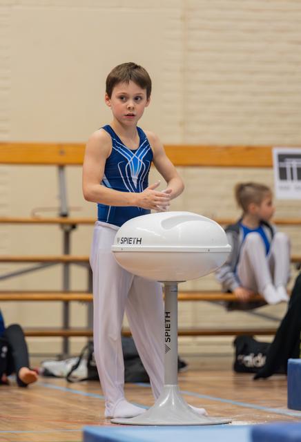 Young male gymnast in blue leotard chalks his hands at a Spieth pommel horse, expression focused and determined.