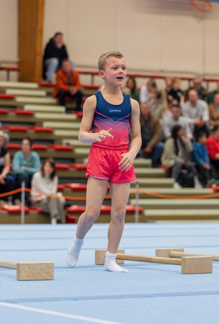 Young boy in blue and pink Quatro leotard performs floor routine, expression intense, audience watching in bleachers behind.