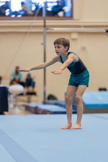 Young gymnast in teal leotard stands focused on floor mat, arms extended forward in a determined ready pose.