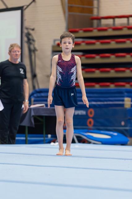 Young male gymnast stands barefoot on the floor mat, composed and focused, with a coach observing in the background.