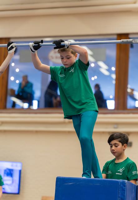 Focused boy in green grips horizontal bar standing on blue block, younger teammate watches attentively below.