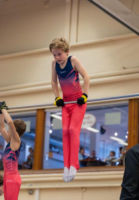 Young boy in red and navy leotard grips the horizontal bar with intense focus during a gymnastics event.