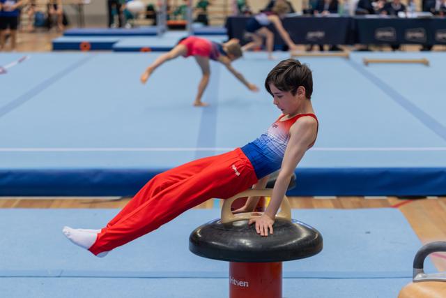 Young male gymnast holds a tuck position on a mushroom pommel, face intent and focused, in a busy competition gym.