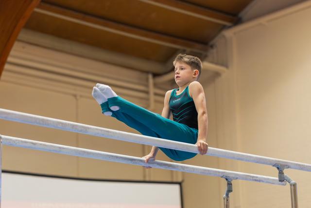 Focused young boy performing a leg raise on parallel bars, wearing a teal leotard, in an indoor gymnastics hall.