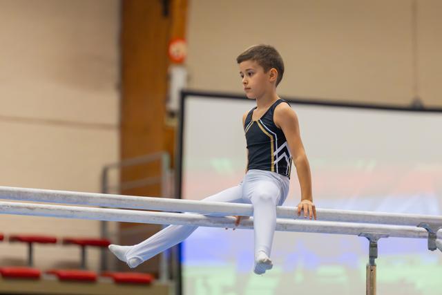 Young boy gymnast sits poised on parallel bars, legs extended, expression focused and calm in a gymnasium setting.