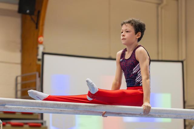 Focused young male gymnast performs a straddle hold on parallel bars, gazing ahead with calm determination in a sports hall.