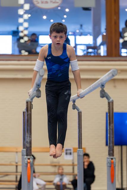Young boy gymnast holds himself up on parallel bars with focused concentration, wearing blue GK leotard in gymnasium.