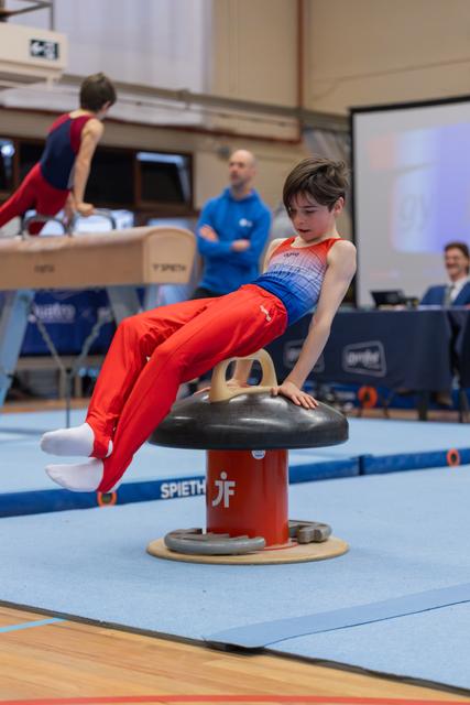 Young male gymnast performs focused pommel horse routine, legs extended, in a blue gymnasium with coach watching behind.