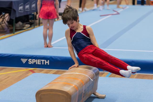 Young boy in red leotard concentrates intensely while performing a pommel horse routine at an indoor gymnastics meet.