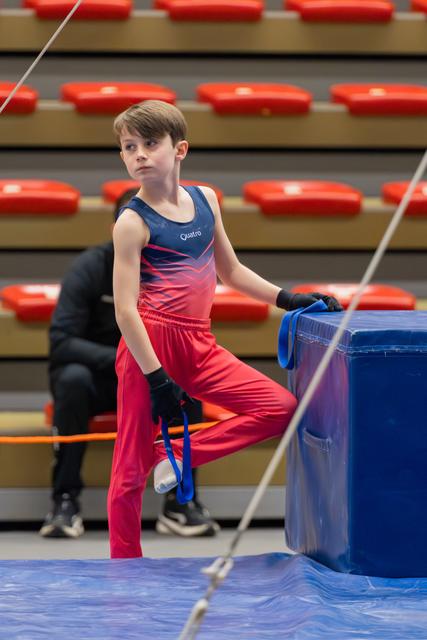 Young male gymnast in red and navy Quatro leotard rests a leg on a blue vault, looking focused and composed.