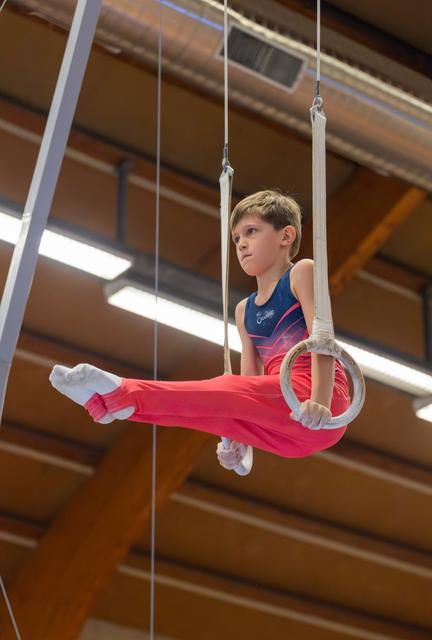 Determined young boy holds gymnastics rings in a split position, legs extended, concentrated gaze in an indoor gym.