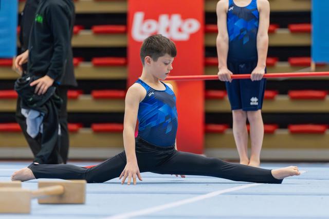 Focused young male gymnast performs a side split on the floor mat, eyes downcast, during a gymnastics event.